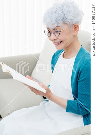 A senior woman sitting on a sofa reading a book. 140957137