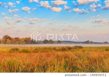 Summer landscape with green misty meadow, trees and sky. Fog on the grassland Summer landscape with green misty meadow, trees and sky. Fog on the grassland 140959351
