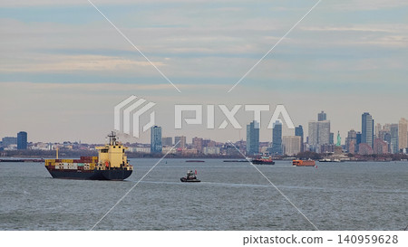 Horizontal wide shot of a loaded container ship and a small tugboat sailing in New York harbor with the city skyline and Statue of Liberty visible far away Horizontal wide shot of a loaded container ship and a small tugboat sailing in New York harbor with the city skyline and Statue of Liberty visible far away 140959628