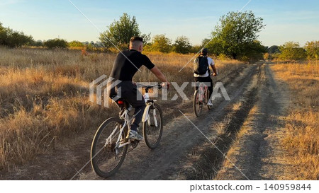 Young male friends ride mountain ski bikes along trail on sunny summer day. Beautiful golden sunlight at background. Concept of active lifestyle, adventure spirit and carefree weekend outdoor Young male friends ride mountain ski bikes along trail on sunny summer day. Beautiful golden sunlight at background. Concept of active lifestyle, adventure spirit and carefree weekend outdoor 140959844