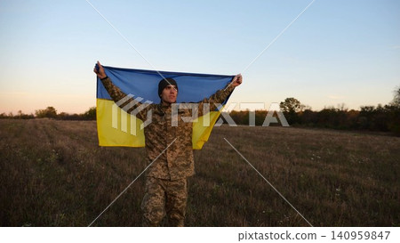 Young male military in uniform standing with raised flag Ukraine above his head at field. Soldier of ukrainian army lifted blue-yellow banner as symbol of victory against russian aggression on field Young male military in uniform standing with raised flag Ukraine above his head at field. Soldier of ukrainian army lifted blue-yellow banner as symbol of victory against russian aggression on field 140959847