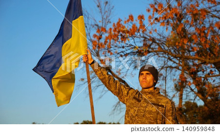 Male ukrainian army soldier lifting blue-yellow banner in honor of victory against russian aggression. Young military in camouflage uniform raised flag of Ukraine at countryside. Invasion resistance Male ukrainian army soldier lifting blue-yellow banner in honor of victory against russian aggression. Young military in camouflage uniform raised flag of Ukraine at countryside. Invasion resistance 140959848