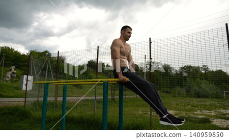 Strong man trains abdominal muscles raising legs on parallel bars at sports ground. Young shirtless sportsman exercising at nature. Sporty athlete working out at nature. Concept of sportive lifestyle Strong man trains abdominal muscles raising legs on parallel bars at sports ground. Young shirtless sportsman exercising at nature. Sporty athlete working out at nature. Concept of sportive lifestyle 140959850