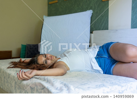 Young woman lying comfortably on her back in bed, dressed in casual shorts and a white top, finding comfort and tranquility during a lazy day at home 140959869