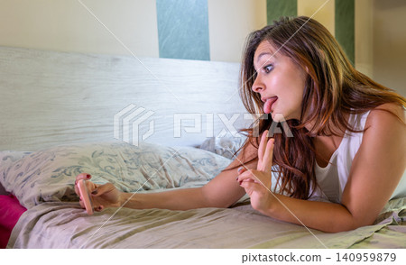 Young woman lying on bed making a silly face with tongue out and a peace sign gesture while taking a selfie with her smartphone, showing playfulness and self expression Young woman lying on bed making a silly face with tongue out and a peace sign gesture while taking a selfie with her smartphone, showing playfulness and self expression 140959879