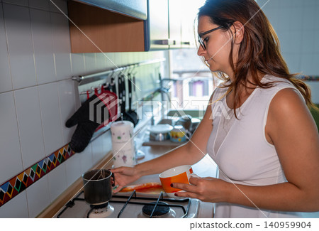 Woman standing in a home kitchen, preparing a hot drink on the gas stove, holding a cup and getting ready to pour while starting her morning routine 140959904