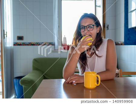 Young woman wearing glasses sitting at kitchen table, enjoying a fresh yellow apple and a drink from a yellow mug, embracing a healthy lifestyle indoors Young woman wearing glasses sitting at kitchen table, enjoying a fresh yellow apple and a drink from a yellow mug, embracing a healthy lifestyle indoors 140959908