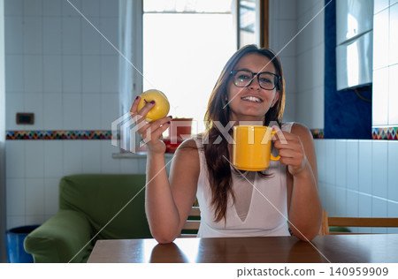 Young woman wearing glasses sitting at a table in a kitchen, holding a yellow apple and a yellow mug, smiling, embodying a healthy lifestyle and morning routine Young woman wearing glasses sitting at a table in a kitchen, holding a yellow apple and a yellow mug, smiling, embodying a healthy lifestyle and morning routine 140959909