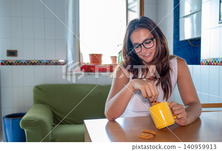Young woman wearing glasses and a white top, smiling while dipping a cookie into her yellow coffee cup, enjoying a relaxed breakfast moment at a wooden table in her home kitchen Young woman wearing glasses and a white top, smiling while dipping a cookie into her yellow coffee cup, enjoying a relaxed breakfast moment at a wooden table in her home kitchen 140959913