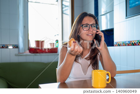 Young woman wearing glasses looking confused and holding a pastry while having a serious conversation on a mobile phone at a kitchen table Young woman wearing glasses looking confused and holding a pastry while having a serious conversation on a mobile phone at a kitchen table 140960049