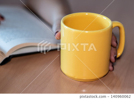 Woman's hand holding a prominent yellow ceramic mug on a wooden desk, while her other hand is softly holding an open book, creating a scene of relaxation and learning Woman's hand holding a prominent yellow ceramic mug on a wooden desk, while her other hand is softly holding an open book, creating a scene of relaxation and learning 140960062
