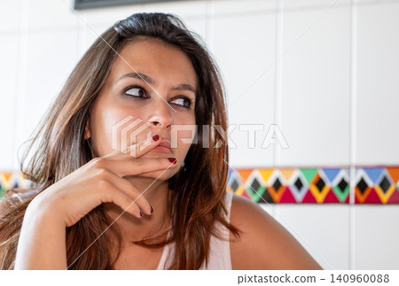 Young brunette woman looking away in deep thought, concentrating on a serious problem while touching her lips in a pensive gesture against a tiled background Young brunette woman looking away in deep thought, concentrating on a serious problem while touching her lips in a pensive gesture against a tiled background 140960088