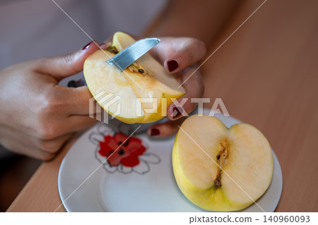 Female hands holding a quartered apple, using a small knife to remove the core and seeds, preparing a healthy fruit snack on a plate with another apple half nearby Female hands holding a quartered apple, using a small knife to remove the core and seeds, preparing a healthy fruit snack on a plate with another apple half nearby 140960093