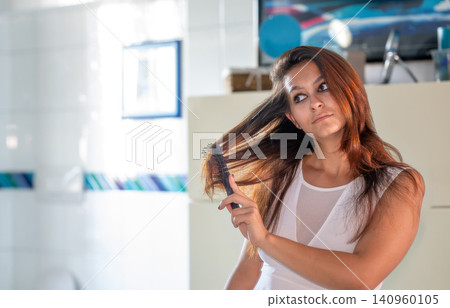 Young woman standing in a bathroom and combing her long brown hair, preparing for the day or evening with a focus on beauty and self care routine Young woman standing in a bathroom and combing her long brown hair, preparing for the day or evening with a focus on beauty and self care routine 140960105