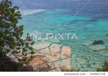 Clear turquoise water with visible rocks and shoreline vegetation along a coastal area under a bright sky Clear turquoise water with visible rocks and shoreline vegetation along a coastal area under a bright sky 140962336