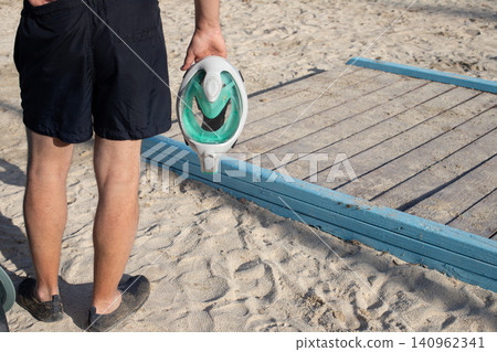 Man holding a green and white beach toy while standing on sandy shore near wooden walkway leading to the water Man holding a green and white beach toy while standing on sandy shore near wooden walkway leading to the water 140962341