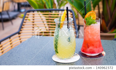 Two colorful cocktails on a table outdoors, one with a pineapple slice and the other garnished with an orange slice and mint leaves Two colorful cocktails on a table outdoors, one with a pineapple slice and the other garnished with an orange slice and mint leaves 140962372