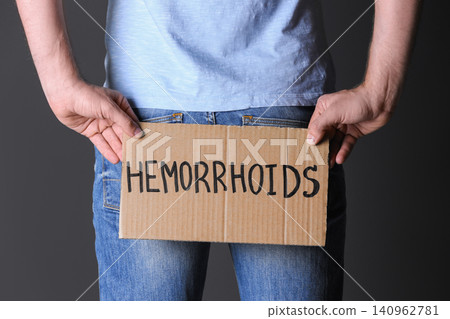 Man holding carton sign with word HEMORRHOIDS on black background, closeup Man holding carton sign with word HEMORRHOIDS on black background, closeup 140962781