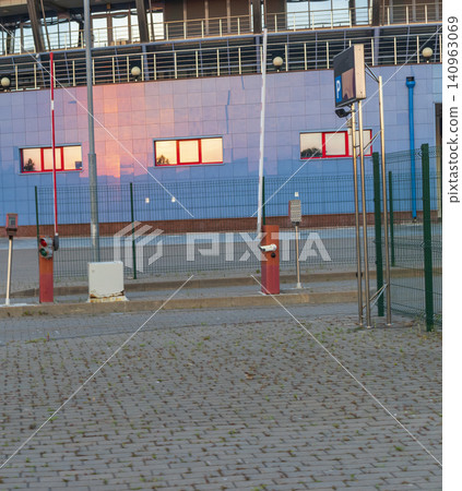 Automated parking lot entrance with barrier gates, security ticket terminal, and a wire mesh safety fence outside a modern stadium building. 140963069