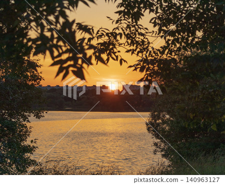 Beautiful golden hour sunset over a peaceful lake with a city building skyline on the horizon framed by dark tree leaves. Nature Beautiful golden hour sunset over a peaceful lake with a city building skyline on the horizon framed by dark tree leaves. Nature 140963127