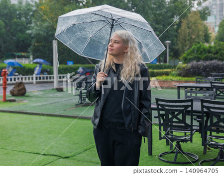 Young woman with blonde hair stands under a clear umbrella during a daytime rain shower Young woman with blonde hair stands under a clear umbrella during a daytime rain shower 140964705