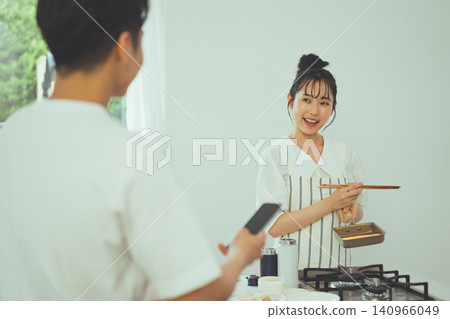 廚房場景:一名女子一邊煮飯一邊微笑著越過一名手持智慧型手機的男子的肩膀與人交談。 廚房場景:一名女子一邊煮飯一邊微笑著越過一名手持智慧型手機的男子的肩膀與人交談。 140966049