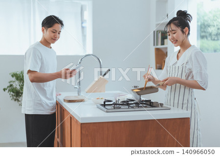 廚房場景:一名男子正在看智慧型手機,一名微笑的女子正在用煎鍋煮飯。 廚房場景:一名男子正在看智慧型手機,一名微笑的女子正在用煎鍋煮飯。 140966050