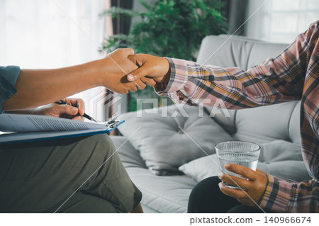 Therapist shakes hands with female client holding water during private counseling on sofa, showing trust, mental health support, professional care, empathy, recovery, and stress relief indoors. Therapist shakes hands with female client holding water during private counseling on sofa, showing trust, mental health support, professional care, empathy, recovery, and stress relief indoors. 140966674