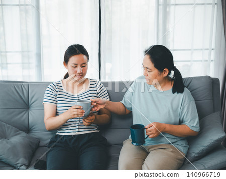 Asian mother giving cup of tea to adult daughter on sofa at home, concept of family support, caregiving, emotional comfort, recovery, kindness, and home wellness. 140966719