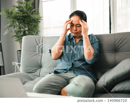 Middle aged Asian woman sitting on sofa with laptop and touching temples from headache, stress, migraine, eye strain, burnout, or work from home pressure. Middle aged Asian woman sitting on sofa with laptop and touching temples from headache, stress, migraine, eye strain, burnout, or work from home pressure. 140966720