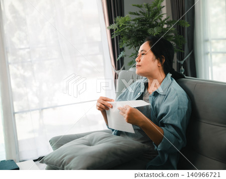 Asian woman sitting on sofa at home holding white envelope and thinking calmly near window, concept of personal finance, letter, notification, insurance, or life decision. 140966721