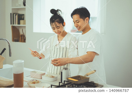 A couple smiling and making bento boxes side-by-side in the kitchen, along with an egg roll pan. A couple smiling and making bento boxes side-by-side in the kitchen, along with an egg roll pan. 140966787