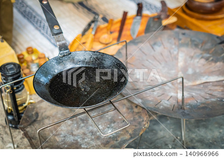 A close-up of a well-used black cast iron frying pan set on a campfire stand. 140966966