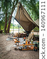 A rugged pup tent site set up by the water in a beautiful forest, with sunlight filtering through the trees. 140967111