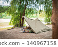 A military pup tent blends seamlessly into the tranquil waterside forest, alongside stylish camping gear. 140967158