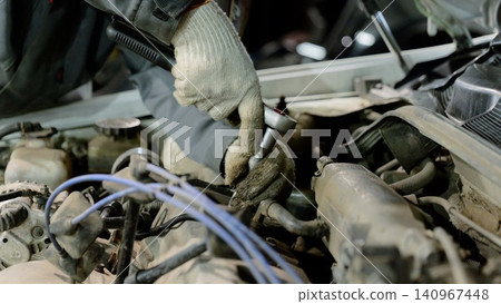 Car mechanic wearing gloves is using a ratchet to skillfully repair the engine of a vehicle with the hood raised, surrounded by tools in a busy garage workshop 140967448