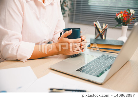 Professional holding coffee beside laptop on office desk with reports and stationery, ideal for business productivity, remote work, planning, corporate lifestyle, online workflow, and workspace. Professional holding coffee beside laptop on office desk with reports and stationery, ideal for business productivity, remote work, planning, corporate lifestyle, online workflow, and workspace. 140968097