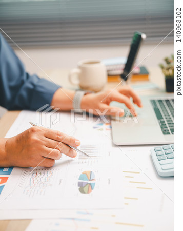 Professional reviewing financial charts with pen on office desk beside laptop and calculator, ideal for accounting analysis, budget planning, audit, corporate reporting, and data strategy. 140968102