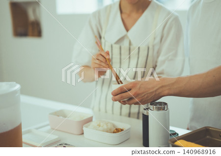 A close-up shot of a woman's hands (faceless) as she carefully packs side dishes into a bento box using chopsticks in the kitchen. A close-up shot of a woman's hands (faceless) as she carefully packs side dishes into a bento box using chopsticks in the kitchen. 140968916