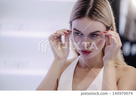 Young woman trying on eyeglasses in optical store, happy customer shopping for new eyewear Young woman trying on eyeglasses in optical store, happy customer shopping for new eyewear 140970074