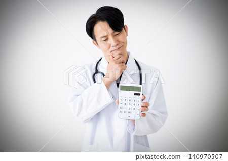 A middle-aged male doctor in a white coat, pondering with a calculator in his hand. A middle-aged male doctor in a white coat, pondering with a calculator in his hand. 140970507