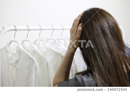 一位女士背對著鏡頭,凝視著衣架上整齊排列的衣服,陷入沉思。 一位女士背對著鏡頭,凝視著衣架上整齊排列的衣服,陷入沉思。 140972504