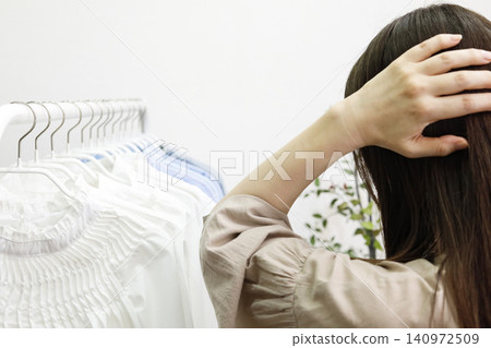 The back view of a woman looking at clothes lined up on a hanger rack, deep in thought. The back view of a woman looking at clothes lined up on a hanger rack, deep in thought. 140972509