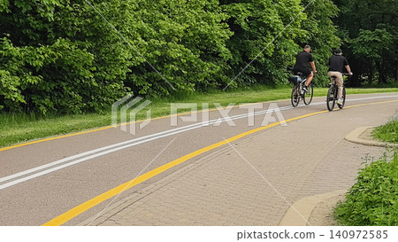 Two cyclists riding away on a marked bike lane through a lush green park 140972585