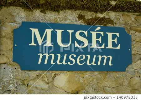 Museum sign displayed in French and English on a traditional stone wall. Bilingual directional signage for cultural heritage and tourism in a historic setting. Museum sign displayed in French and English on a traditional stone wall. Bilingual directional signage for cultural heritage and tourism in a historic setting. 140978013