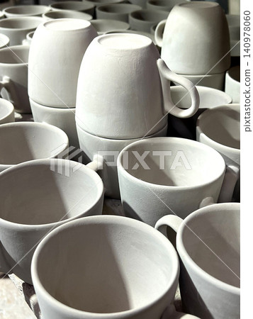 Ceramic cups and mugs crafted from clay are neatly arranged in a pottery studio, showcasing their unfinished, raw form in natural light 140978060