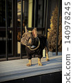 A fashionable dog wearing a black sweater and chain necklace stands proudly on a balcony railing against an urban backdrop at sunset 140978245