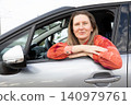 Driver woman with blonde hair sits in car with arms resting outside window. She smiles while parked in urban area during daytime. Horizontal. 140979761