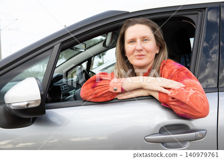 Driver woman with blonde hair sits in car with arms resting outside window. She smiles while parked in urban area during daytime. Horizontal. Driver woman with blonde hair sits in car with arms resting outside window. She smiles while parked in urban area during daytime. Horizontal. 140979761