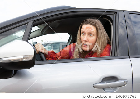 Driver blonde woman looks surprised in car looking outside in parking lot during daytime. Horizontal. Driver blonde woman looks surprised in car looking outside in parking lot during daytime. Horizontal. 140979765